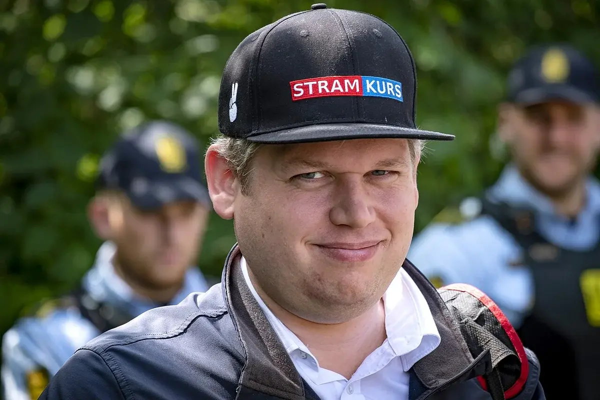 Rasmus Paludan wearing a Stram Kurs cap, standing with police nearby.