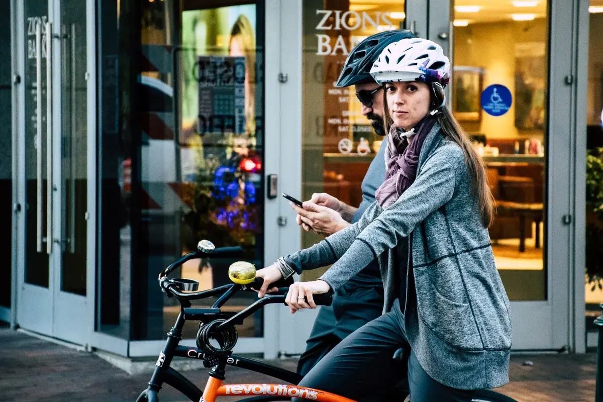 A man and woman wearing helmets stand with bicycles outside a bank while the man checks his phone.