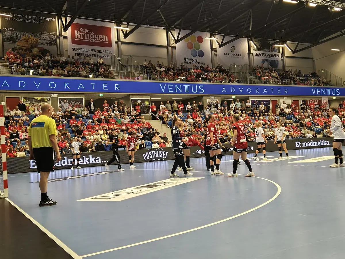 Women’s handball match in an indoor arena with players clustered near the goal and a referee watching.