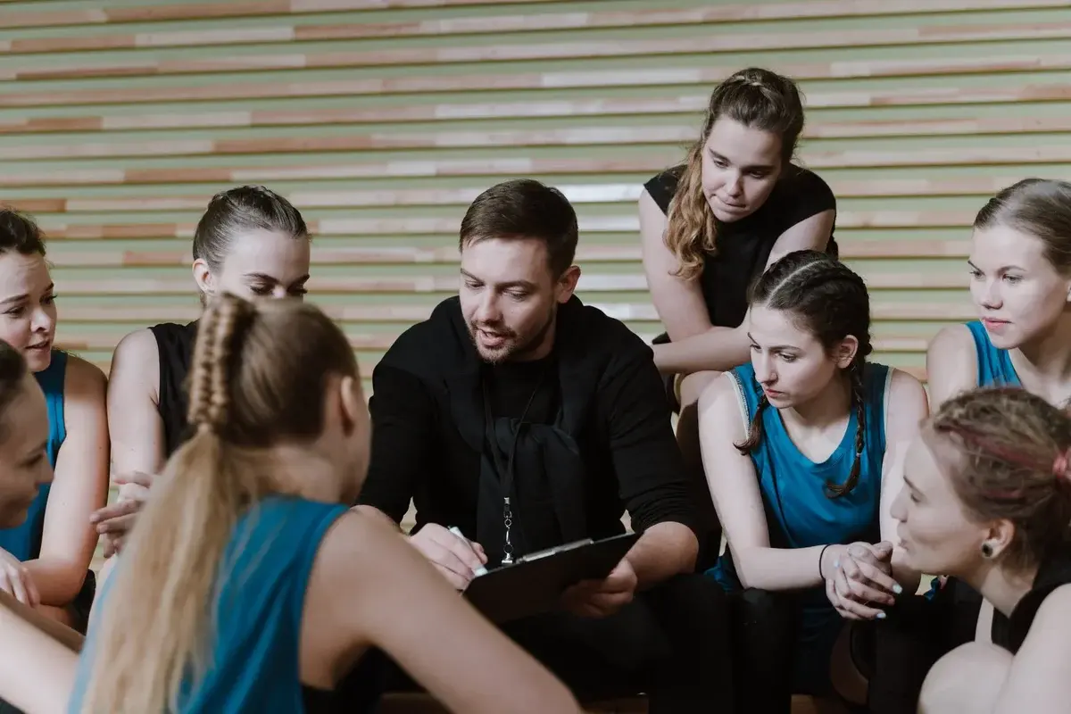 A coach talks to a women’s sports team in a gym.