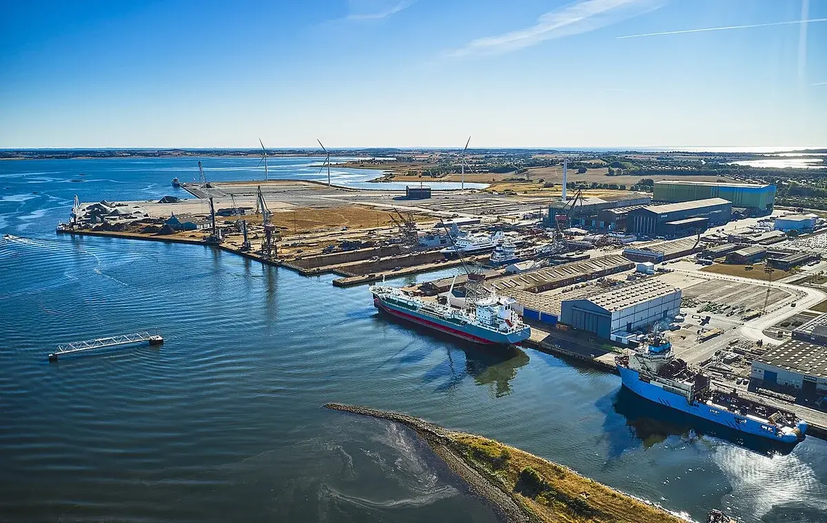 Aerial view of an industrial harbor with ships, cranes, and wind turbines.