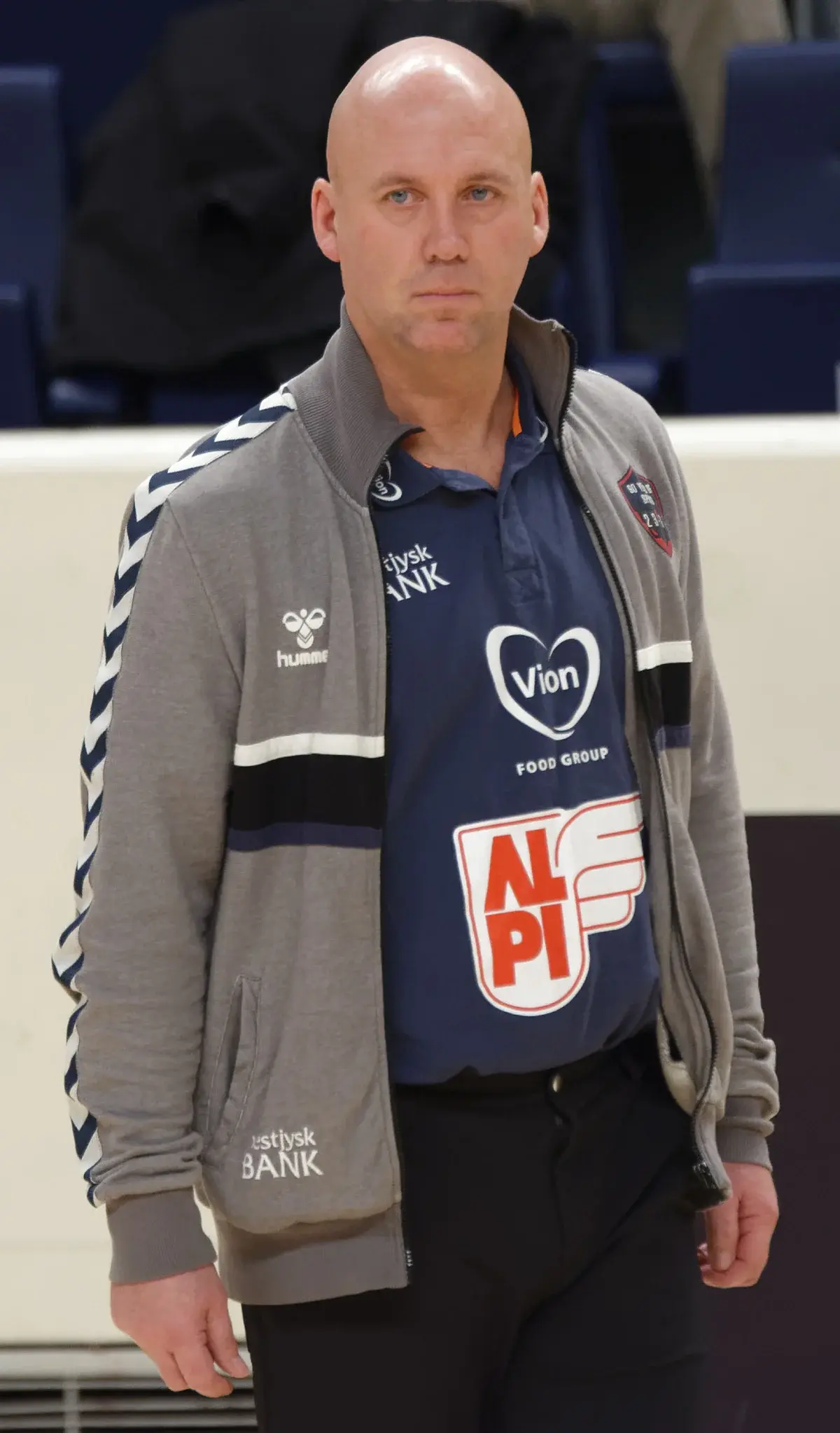 A bald man in a gray jacket stands on the sideline at an indoor handball arena.