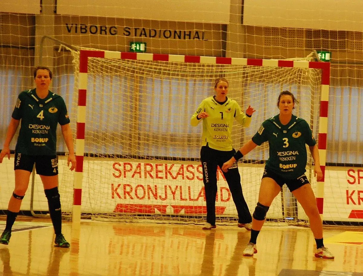 Three Viborg HK handball players defending in front of the goal in an indoor arena.